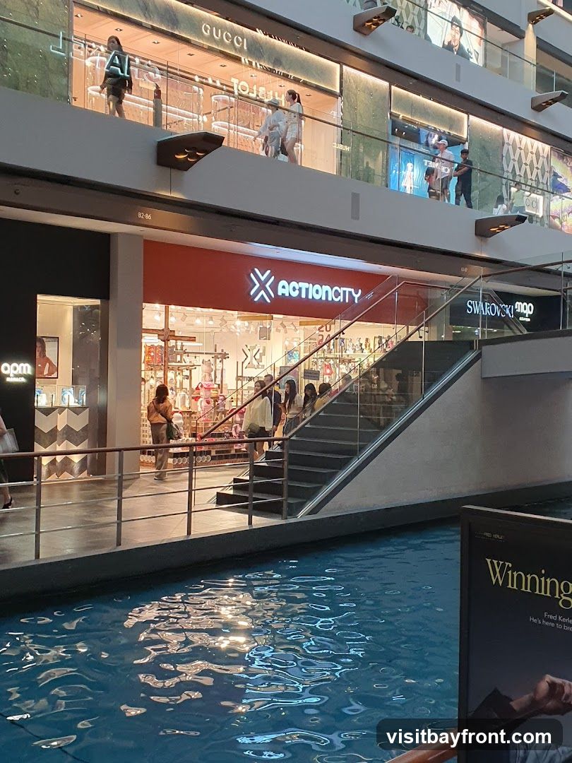 A view of the ACTIONCITY store entrance in a mall, with a canal in the foreground. The store has a bright orange facade with the store name in white lettering. Above ACTIONCITY, the GUCCI store is visible. Other stores like Swarovski and apm are also present. People are seen walking on the escalators and inside the stores. The water in the canal reflects the lights from the mall.