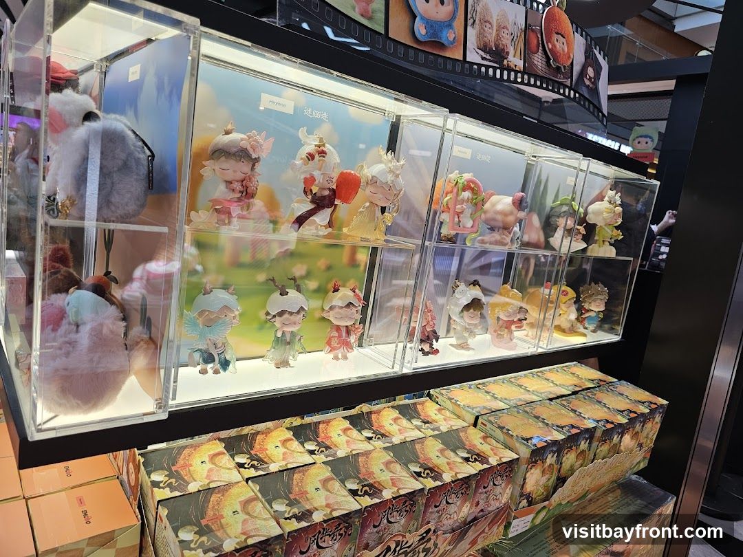 A retail display featuring several clear acrylic cases filled with collectible figurines. The figurines are varied in design, with some appearing to be characters from animated series or games, often depicted in whimsical poses. Some cases also contain plush toys. Below the display cases, stacks of boxed merchandise are visible, featuring colorful artwork. The overall impression is of a store selling toys or collectibles.