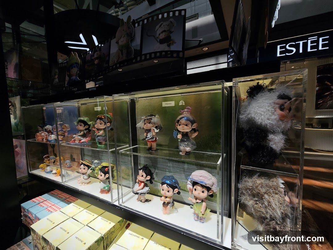 A retail display featuring various collectible figures in clear cases, arranged on shelves. Some figures are depicted in whimsical poses with scenic backgrounds. To the right, larger plush toys are also showcased in display cases. In the background, a sign for 'ESTEE' is visible, suggesting a retail environment. Boxes with branding are stacked in the foreground.
