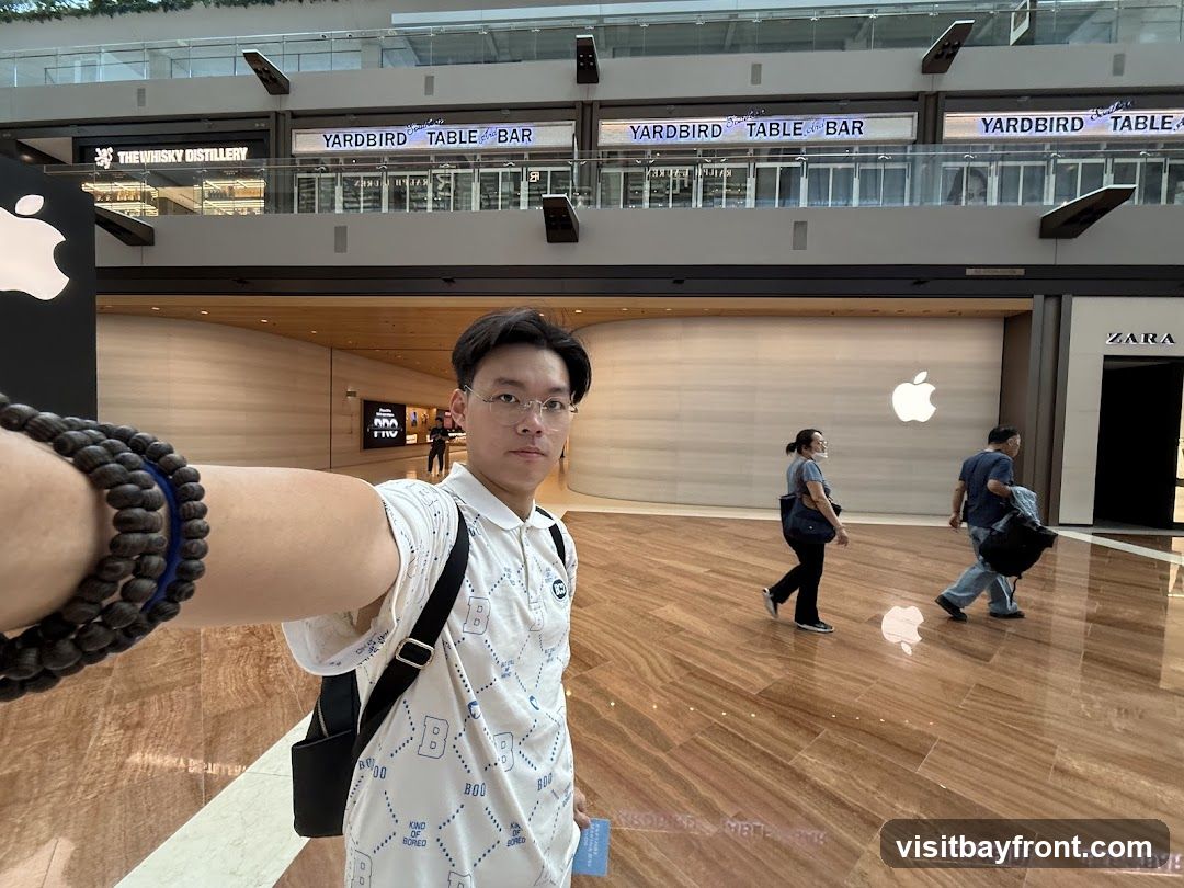 A man takes a selfie in a modern mall. Behind him, the Apple logo is visible on a large white wall. To the right, the ZARA store entrance is seen. In the background, signs for "YARDBIRD TABLE BAR" and "THE WHISKY DISTILLERY" are displayed on an upper level. People are walking in the mall.
