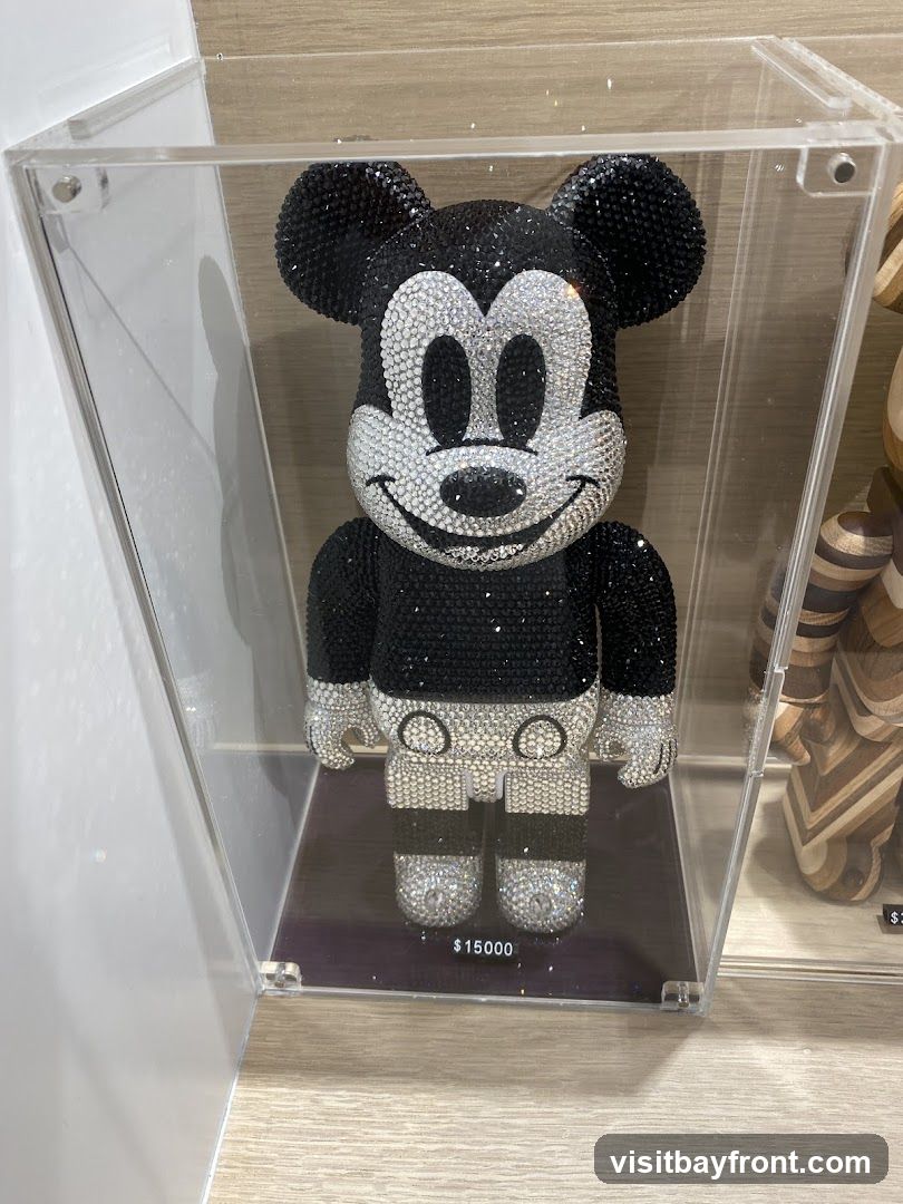 A Mickey Mouse figurine covered in black and white rhinestones is displayed in a clear acrylic case. The figurine is black with a white face, ears, gloves, and shorts. The price tag below reads '$15000'. To the right, another figurine made of wood is partially visible.