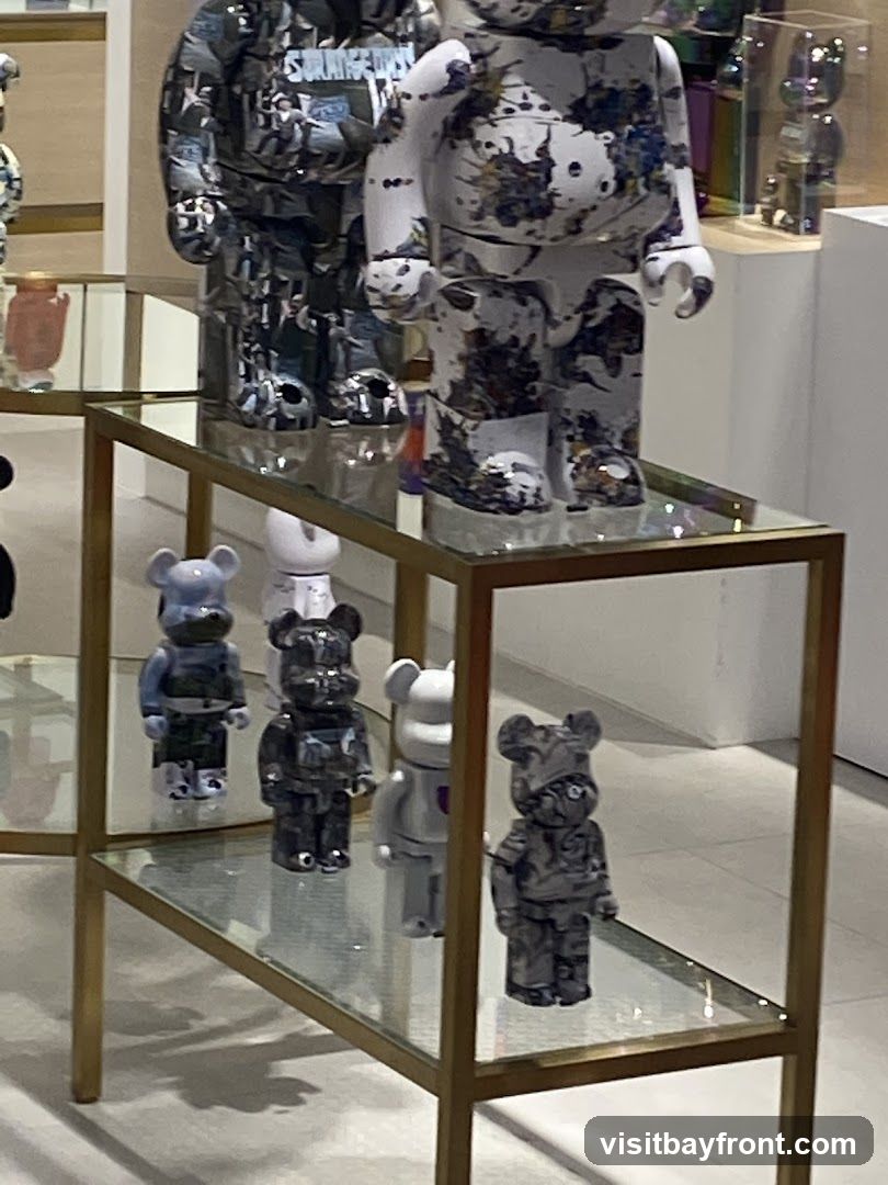 A display shelf with several Bearbrick figurines. The top shelf features two large Bearbricks, one with a black and white collage pattern and the other with a white base splattered with dark paint. The lower shelf holds smaller Bearbricks in various grey and white designs. The figurines are arranged on glass shelves with a gold-colored frame.