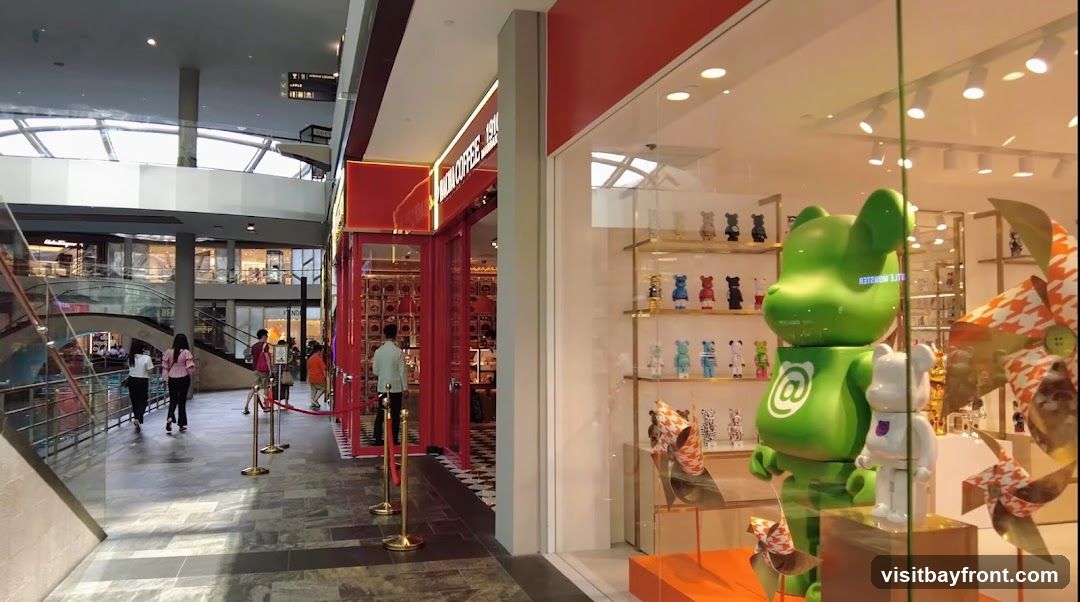 A bright green, large Bearbrick figure with an '@' symbol is displayed prominently in a store window. Next to it, a smaller white Bearbrick figure stands. The window also showcases shelves filled with various smaller Bearbrick figures in different colors and designs. Decorative pinwheels with a houndstooth pattern are also visible. The store appears to be a retail space selling collectible toys or designer merchandise. In the background, a red-accented shop front with the words "INDIAN COFFEE" is visible, suggesting a neighboring establishment.