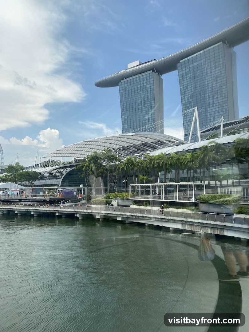 A wide shot of the Marina Bay Sands complex in Singapore, with its iconic three towers connected by a sky-high platform. In the foreground, a body of water reflects the sky and buildings. A promenade runs along the waterfront, with palm trees and modern architecture visible. The sky is blue with scattered white clouds.