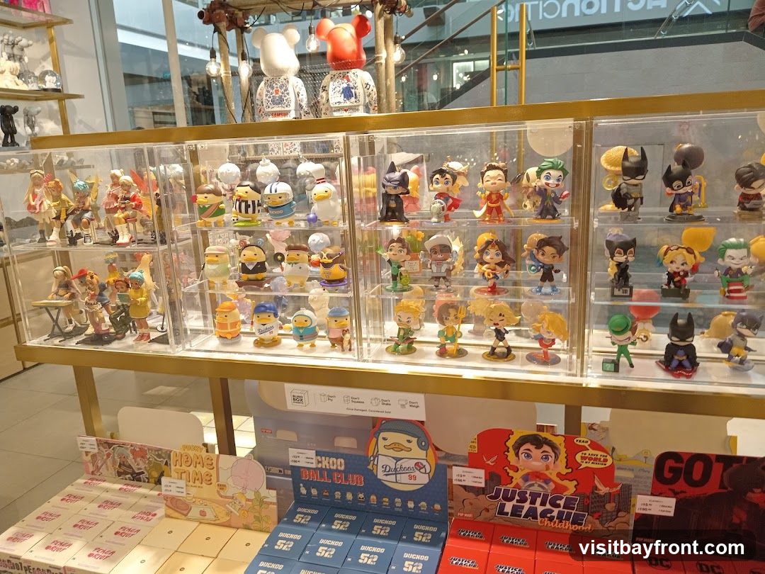 A retail display featuring numerous collectible figurines in clear display cases. The figurines include characters from various franchises such as DC Comics (Batman, Joker, Harley Quinn) and anime-inspired characters. Below the display cases, boxes of collectible toys and merchandise are stacked.