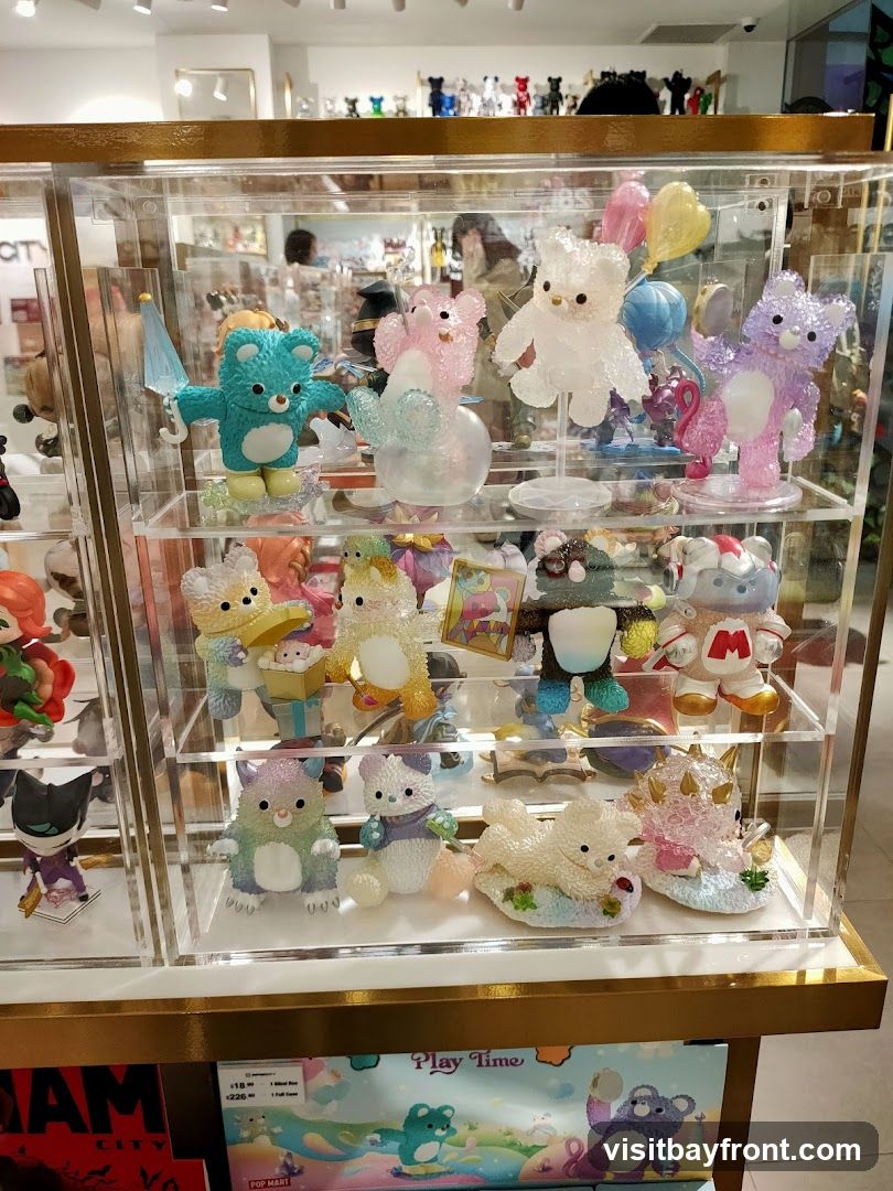 A clear display case filled with various collectible figurines, primarily teddy bear-shaped, in different colors and poses. Some figurines are translucent and sparkly, while others are opaque. They are arranged on multiple shelves within the case. In the background, more figurines are visible on shelves, and a blurred image of people can be seen. Below the display case, a colorful box with cartoon characters and text that includes "Play Time" and pricing information is visible.