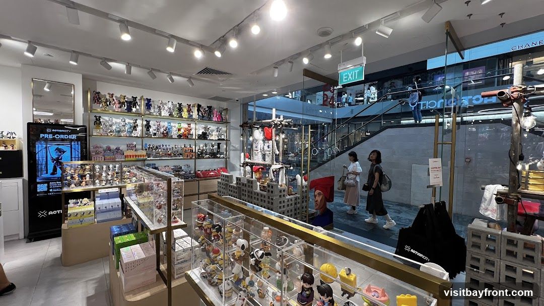 The interior of a store filled with various collectible toys and figures displayed on shelves and in glass cases. A digital screen shows a "PRE-ORDER" advertisement. People are seen browsing the merchandise and walking in the background. An "EXIT" sign is visible above a staircase.