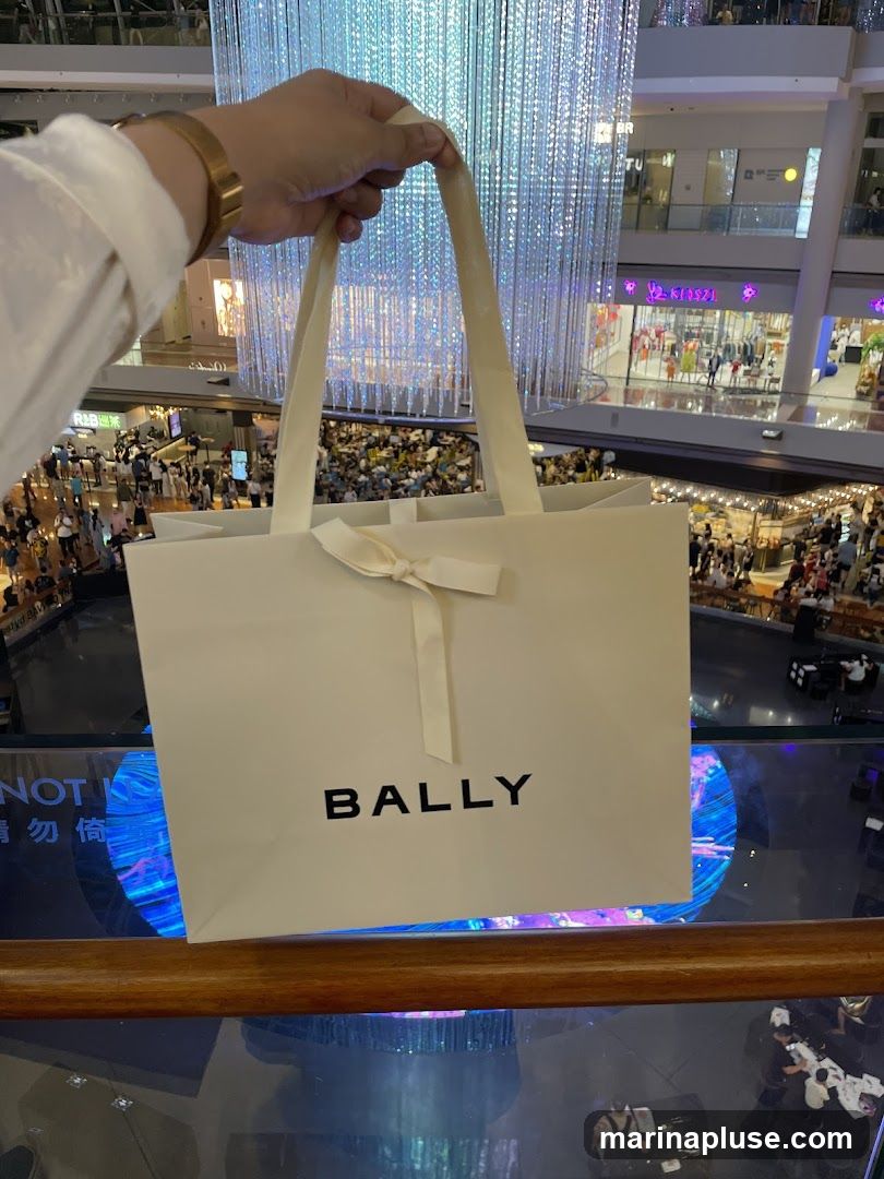 A person holds a white Bally shopping bag in front of a brightly lit, multi-level shopping mall interior. The bag has the Bally logo in black text and a white ribbon tied in a bow. The background shows various shops and a large, illuminated art installation.