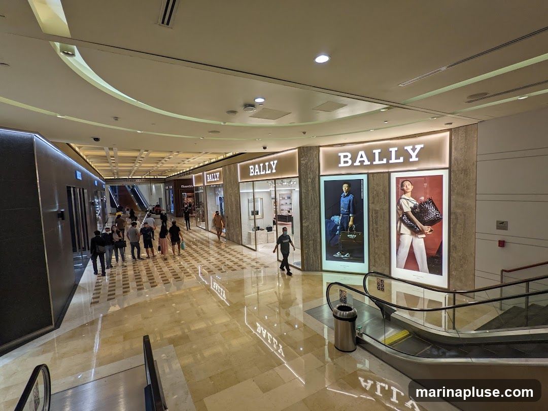 A wide shot of a modern shopping mall interior with several Bally storefronts visible. The stores feature large illuminated signs with the Bally logo and display windows showcasing mannequins and merchandise. Escalators and shoppers are also present in the scene, with polished marble floors reflecting the lights and store displays.