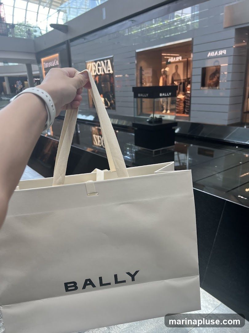A person holds a white Bally shopping bag in front of a row of luxury stores in a mall. The stores visible include Tiffany & Co., Zegna, and Prada. The Bally logo is prominently displayed on the bag.