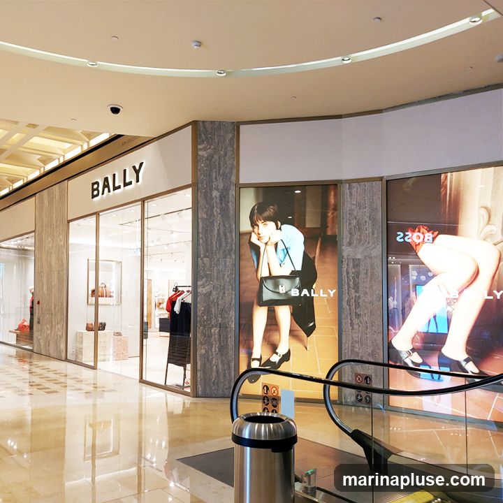 A view of a Bally store entrance in a mall. The storefront features large glass windows displaying merchandise and a prominent Bally logo above the entrance. Adjacent to the store are large advertisements featuring models, one of which also displays the Bally logo. An escalator and a trash can are visible in the foreground.