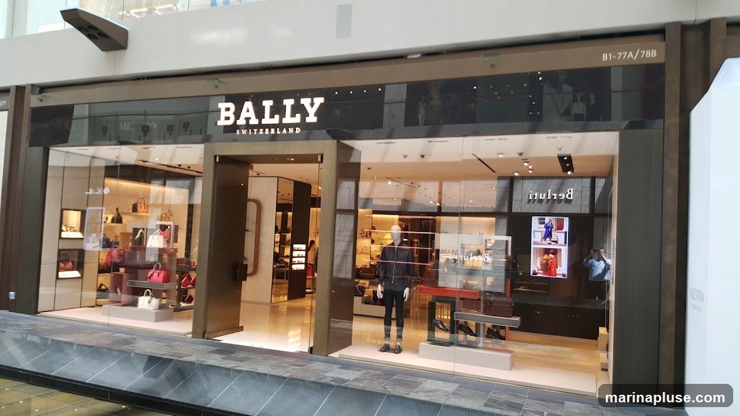 The exterior of a Bally store, a luxury brand, is shown. The store has a large glass facade displaying handbags and accessories. Inside, mannequins are dressed in clothing, and more merchandise is visible on shelves and display units. The Bally logo is prominently displayed above the entrance.