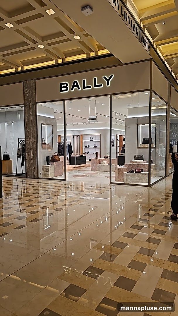 The exterior of a Bally store with its name prominently displayed in illuminated letters above the entrance. The store features large glass windows showcasing handbags and clothing displays inside. The floor in front of the store has a reflective, patterned design.