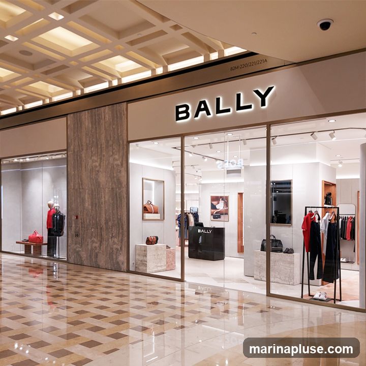 The exterior of a Bally store with a large glass facade. The store's name, "BALLY", is prominently displayed in illuminated white lettering above the entrance. Inside, mannequins display clothing and accessories, and shelves showcase handbags and shoes. The interior is modern and well-lit, with marble-like display stands and polished floors.