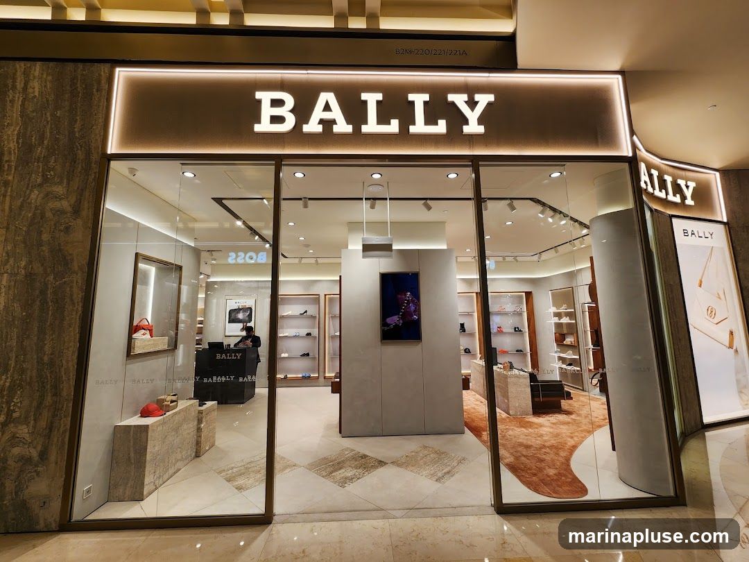 The exterior of a Bally store with large glass windows and doors. The store name "BALLY" is prominently displayed in large white letters above the entrance, illuminated by a soft light. Inside, shelves display shoes and handbags. A person is visible behind a counter. The floor is tiled with a diamond pattern, and a plush orange rug is visible inside.