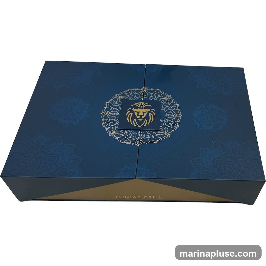 A dark blue gift box with gold accents. The lid is divided into four sections, with a circular gold design in the center featuring a lion's head. The box also has a gold band around the bottom with the text "PUNJAB GRILL" printed on it.