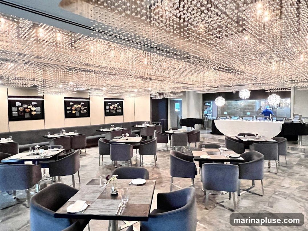A spacious and elegantly decorated restaurant interior is shown. The ceiling is adorned with a multitude of small, sparkling lights, creating a dazzling effect. The dining area features numerous tables set with white tablecloths, cutlery, and glassware, surrounded by plush grey chairs. In the background, a buffet or serving station is visible, with a person in a blue uniform attending to it. The walls are decorated with framed artwork. The overall ambiance suggests a sophisticated dining experience.