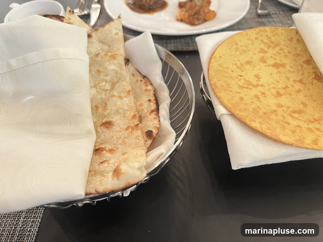 A close-up shot of a bread basket filled with naan and a papadum, both covered with white napkins. In the background, a plate with food is visible.