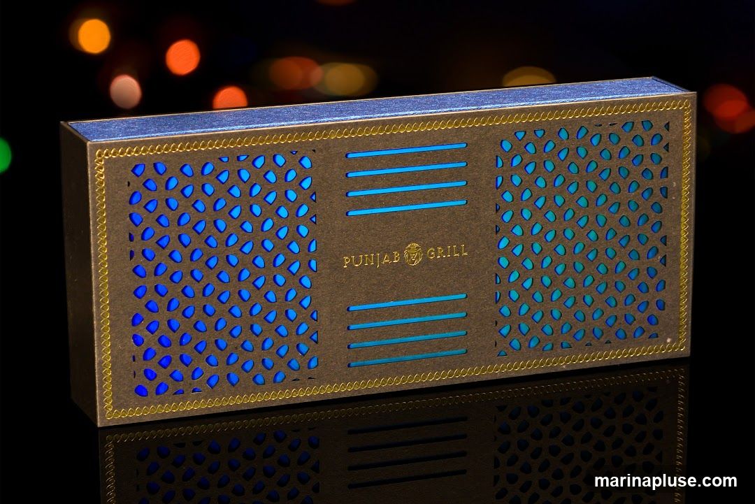 A dark brown rectangular box with intricate cutouts and blue lighting. The box has a gold border and the words "PUNJAB GRILL" are embossed in gold in the center. The background is blurred with bokeh lights in orange, yellow, and green.