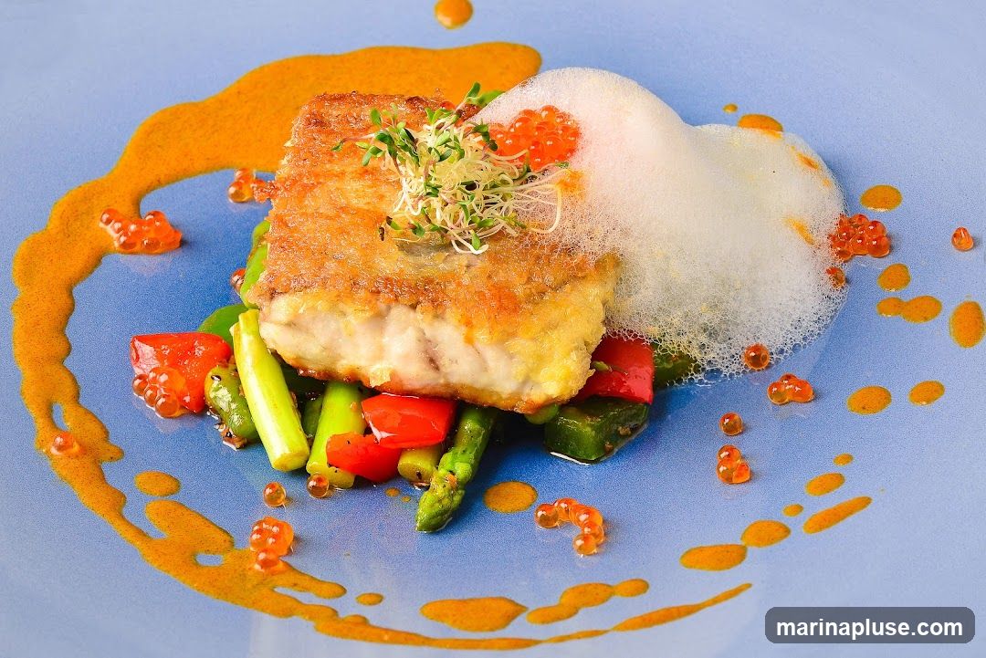 A beautifully plated dish featuring a piece of pan-fried fish with a crispy golden crust, served atop a bed of asparagus and red bell peppers. The dish is garnished with microgreens and a light foam, and adorned with vibrant orange roe and a drizzle of rich, orange-hued sauce. The plate is a soft blue, creating a striking contrast with the colorful food.