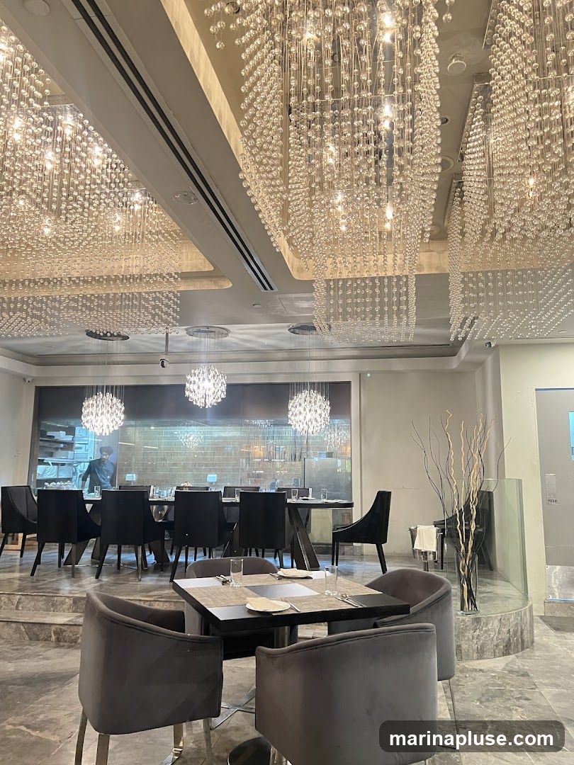 A luxurious dining room interior featuring multiple large, cascading crystal chandeliers that illuminate the space. The room is furnished with plush grey velvet chairs and dark wood tables, set for dining. In the background, a long dining table is visible, with a chef or staff member working in an open kitchen area behind it. A grand piano stands in the corner, adding to the sophisticated ambiance.