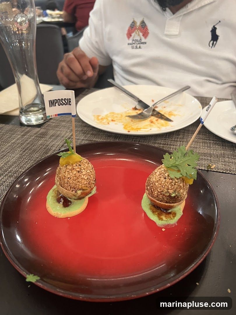 A close-up shot of a red plate with two appetizers. Each appetizer is a spherical item coated in nuts, placed on a small round cracker with a green sauce. A small white flag with the word "IMPOSSIBLE" is stuck into each appetizer. In the background, a person wearing a white polo shirt with American flag patches and a Ralph Lauren logo is visible, along with a partially eaten plate and a tall glass.