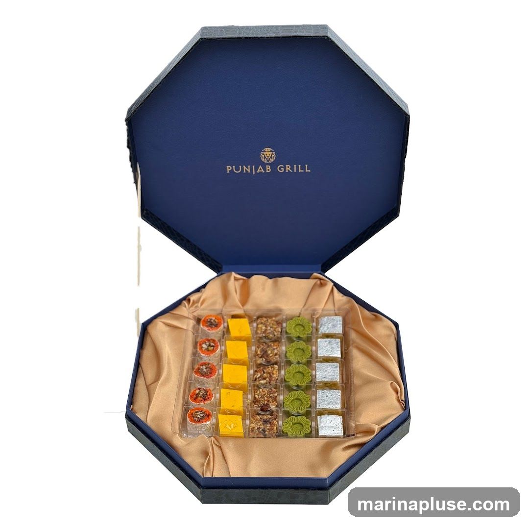 An open, octagonal, dark blue gift box lined with golden satin fabric. Inside, a clear plastic tray holds an assortment of Indian sweets. The sweets are arranged in rows and columns, with distinct types in each column. The lid of the box is open and displays the embossed logo and text 'PUNJAB GRILL'.