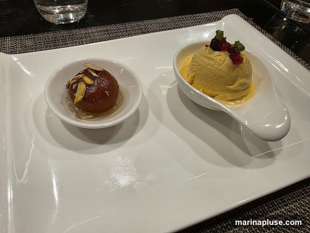 A white platter holds two desserts: a gulab jamun in a small white bowl topped with slivered almonds and pistachios, and a scoop of mango ice cream in a white spoon-shaped dish, garnished with dried berries and mint.