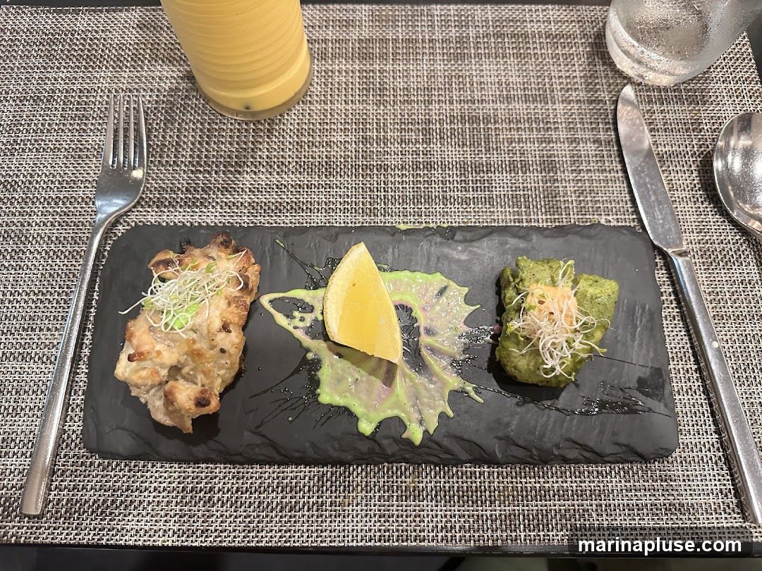 A slate platter holds three small appetizers. The leftmost appetizer appears to be a piece of grilled chicken or fish, garnished with sprouts. In the center is a lemon wedge with a green sauce drizzled around it. The rightmost appetizer is a green patty, possibly a vegetable fritter, also topped with sprouts and a small dollop of sauce. A fork and knife are placed on either side of the platter, and a glass of a yellow beverage is visible in the upper left corner.