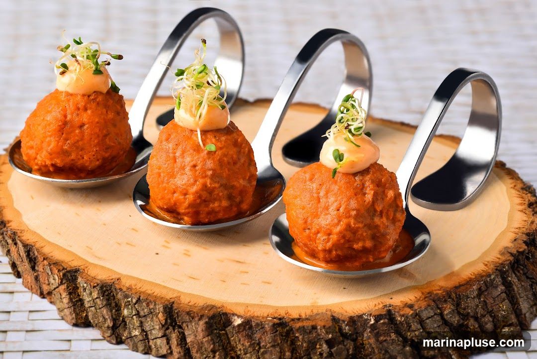 Three appetizer spoons are arranged on a wooden slab. Each spoon holds a round, orange-colored food item topped with a dollop of sauce and sprouts. The spoons have a modern, curved design.