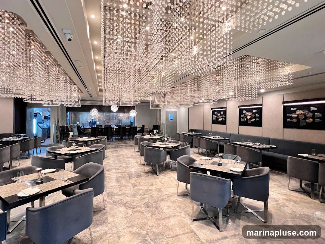 A spacious restaurant dining area with elegant decor. The room features numerous tables and chairs, many with plush grey upholstery. The ceiling is adorned with elaborate, cascading crystal chandeliers that create a dazzling effect. The walls are decorated with framed displays, and a long banquette seating area runs along one side. The floor is a polished marble. In the background, a glimpse of the kitchen area is visible.