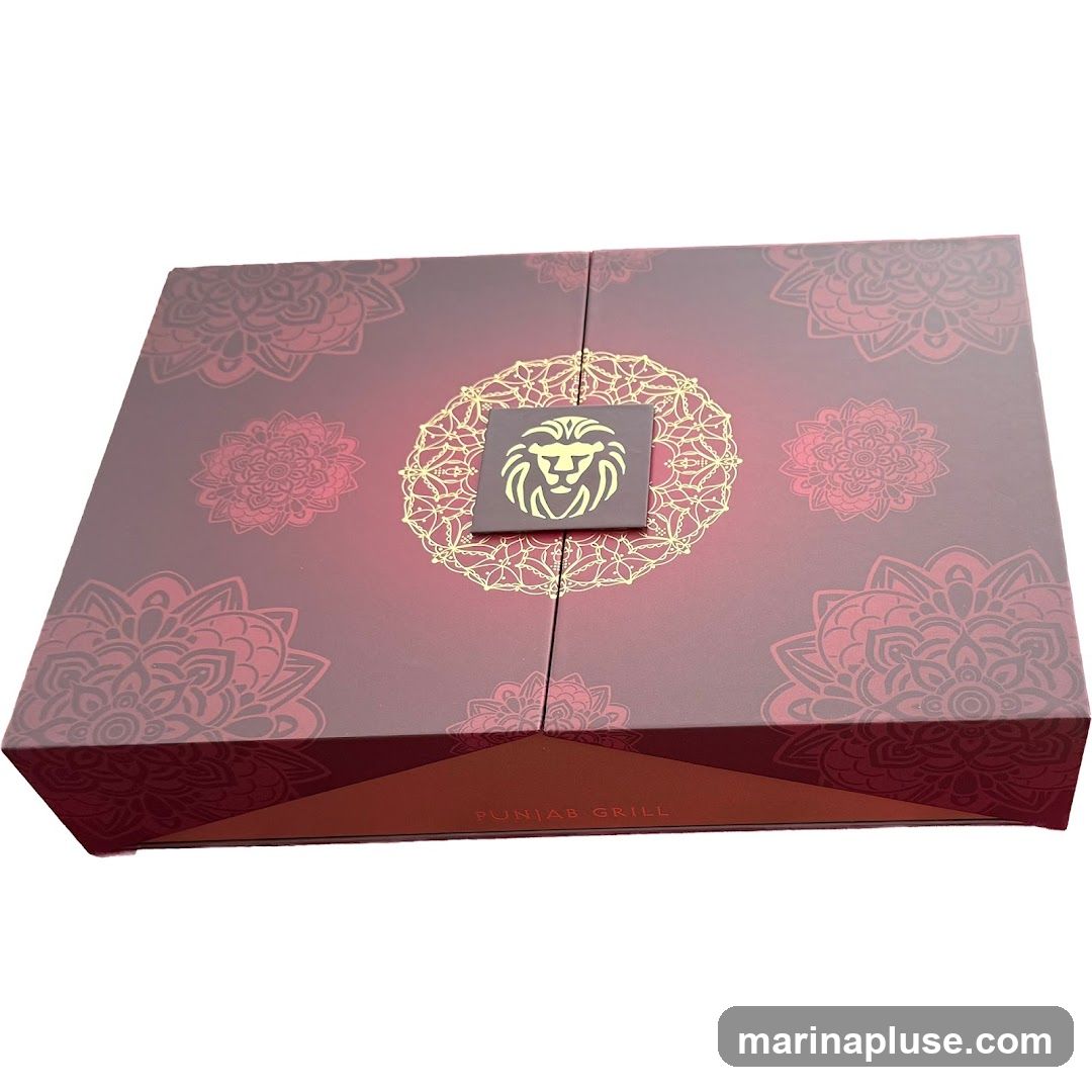 A closed gift box with a maroon and red gradient. The lid is decorated with intricate gold mandala-like patterns and a central square emblem featuring a lion's head. The sides of the box have a similar mandala pattern. The bottom edge of the box has the text "PUNJAB GRILL" printed in gold.