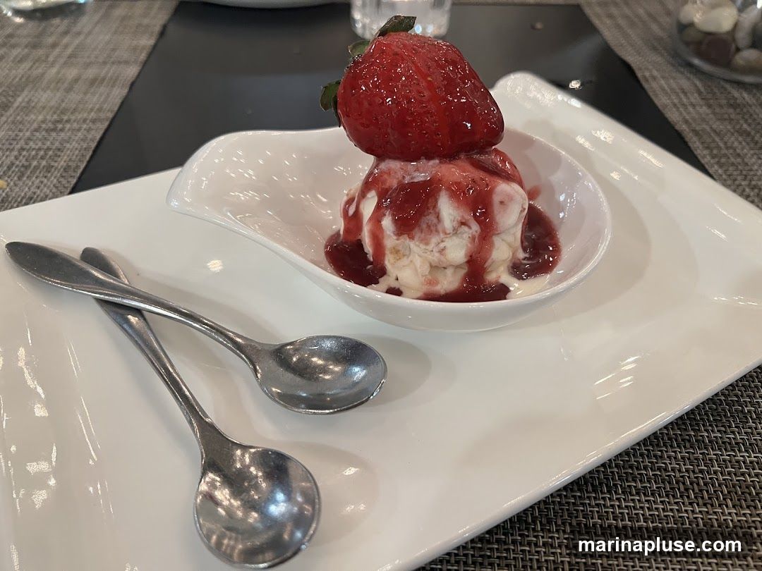 A close-up shot of a dessert featuring two scoops of ice cream in a white bowl, topped with strawberry sauce and a fresh strawberry. Two spoons are placed on a white rectangular plate next to the bowl. The setting appears to be a restaurant table with a textured placemat.