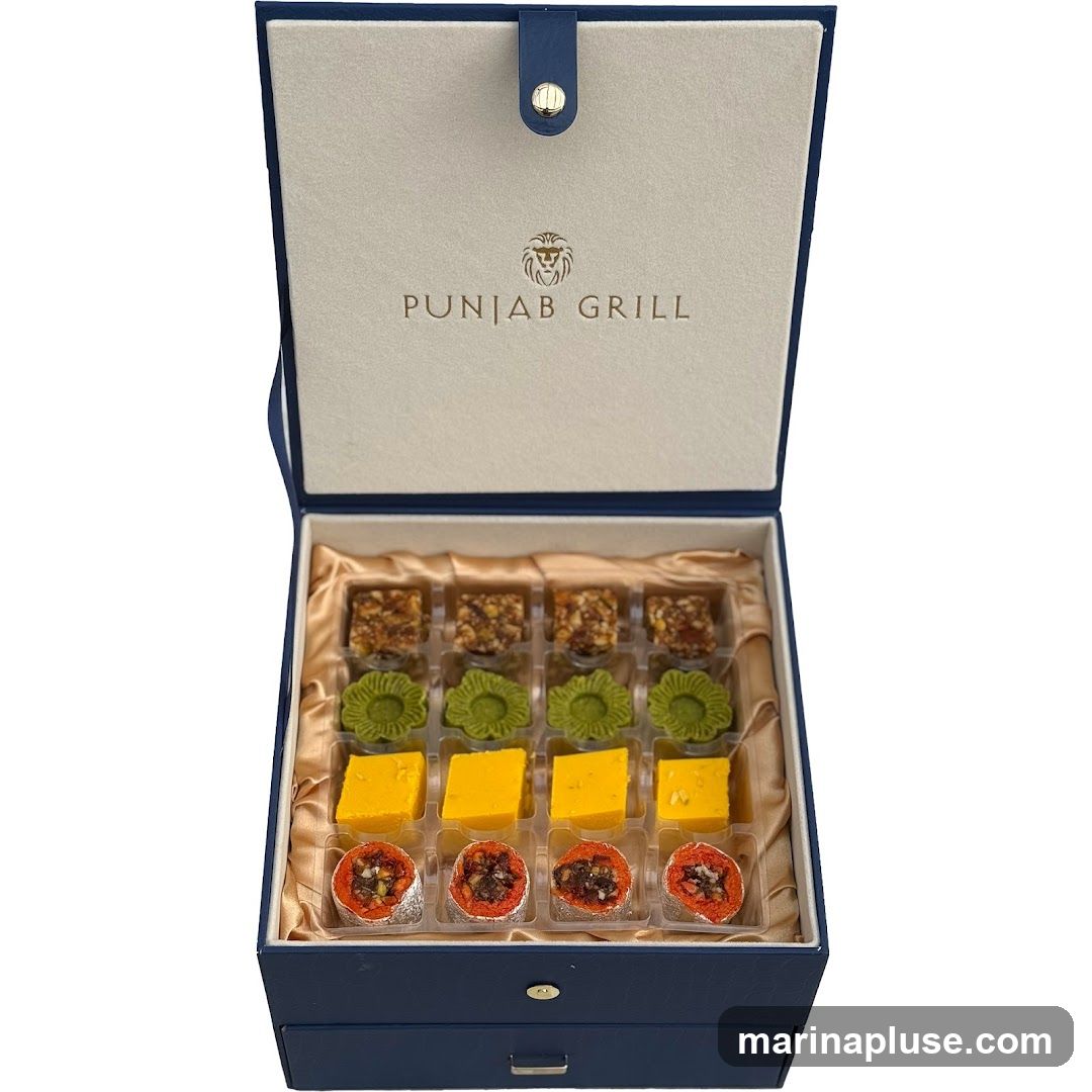 A navy blue gift box is open, revealing an assortment of Indian sweets. The lid of the box is lined with cream-colored fabric and embossed with the logo and name "PUNJAB GRILL". Inside, the sweets are arranged in clear plastic dividers. The top row contains small, square pieces topped with nuts. The second row features green, flower-shaped sweets. The third row has yellow, rectangular sweets. The bottom row contains round, red-orange sweets filled with nuts and dried fruit. The sweets are presented on a golden satin lining.