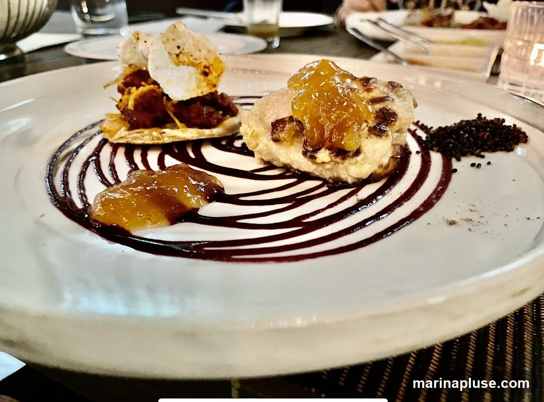 A close-up shot of a dessert on a white plate. The dessert features two distinct components. One is a layered pastry base topped with a dark, savory filling and white meringue shards. The other is a lighter, possibly fried, mound topped with a golden jam. The plate is artistically drizzled with a dark sauce, and a small pile of dark seeds or spices is placed to the right. The setting appears to be a restaurant with soft lighting and other diners in the background.