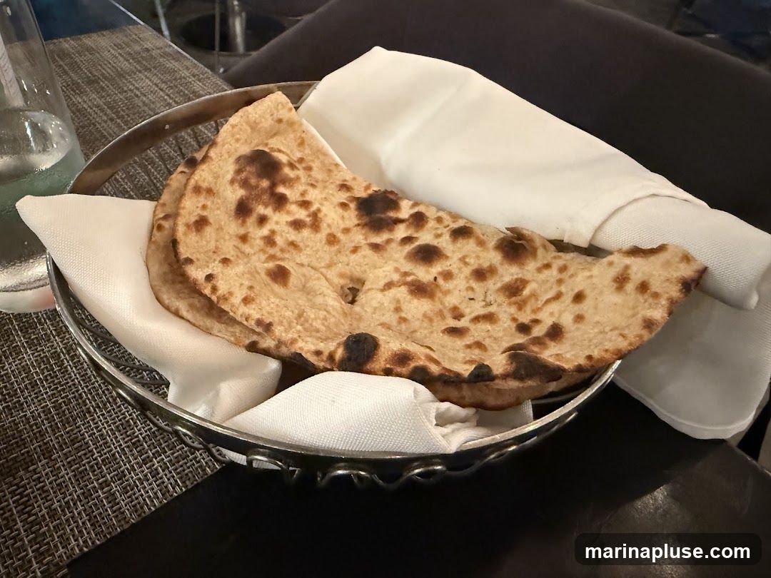 A close-up shot of a metal basket filled with freshly baked naan bread, folded and presented with white cloth napkins. The naan has a slightly charred appearance, indicating it was cooked in a tandoor or similar high-heat oven. A glass of water is visible to the left of the basket.