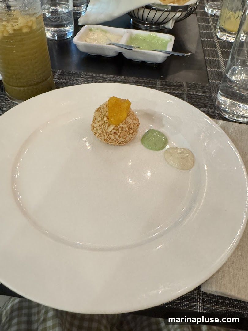A single appetizer, possibly a croquette or arancini, is presented on a white plate. It is coated in what appears to be crushed nuts or seeds and topped with a dollop of orange preserve. Three small puddles of dipping sauces, one green and two white, are arranged beside it. A glass of green beverage, possibly a smoothie or juice, is visible to the left, along with glasses of water and a small dish of condiments in the background. The table is set with a dark placemat.