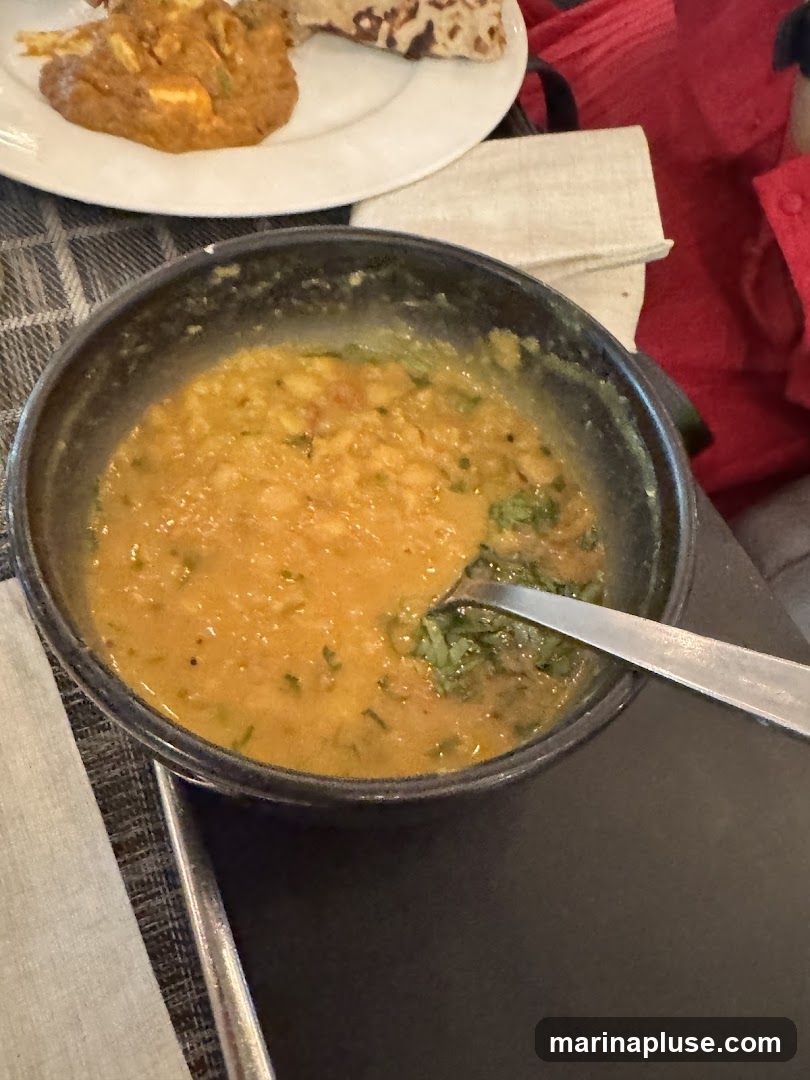 A close-up shot of a bowl of dal, a lentil dish, with a spoon in it. In the background, a white plate holds what appears to be paneer tikka and naan. The food is served on a table with a patterned placemat and a white napkin.