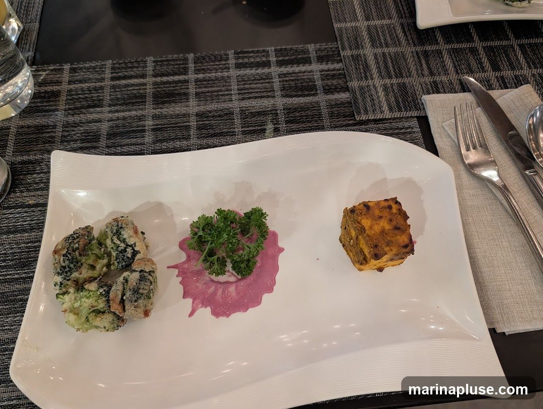 A white rectangular plate holds three appetizers. On the left are several pieces of battered and fried broccoli. In the center is a dollop of pink sauce topped with fresh parsley. On the right is a single cube of tandoori paneer. Cutlery and a glass of water are visible on the table.