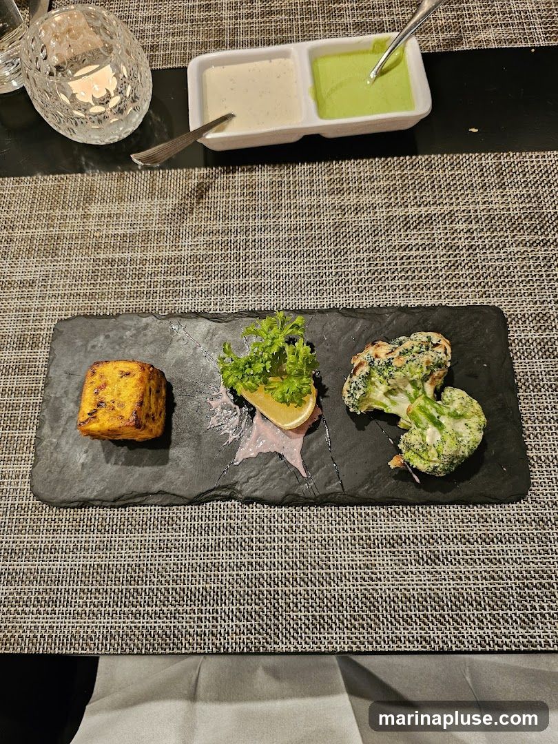 A slate platter holds a square piece of tandoori paneer, a lemon wedge with parsley garnish, and a serving of tandoori broccoli. Two dipping sauces, one white and one green, are in a rectangular dish in the background. A lit candle in a decorative glass holder is to the left.
