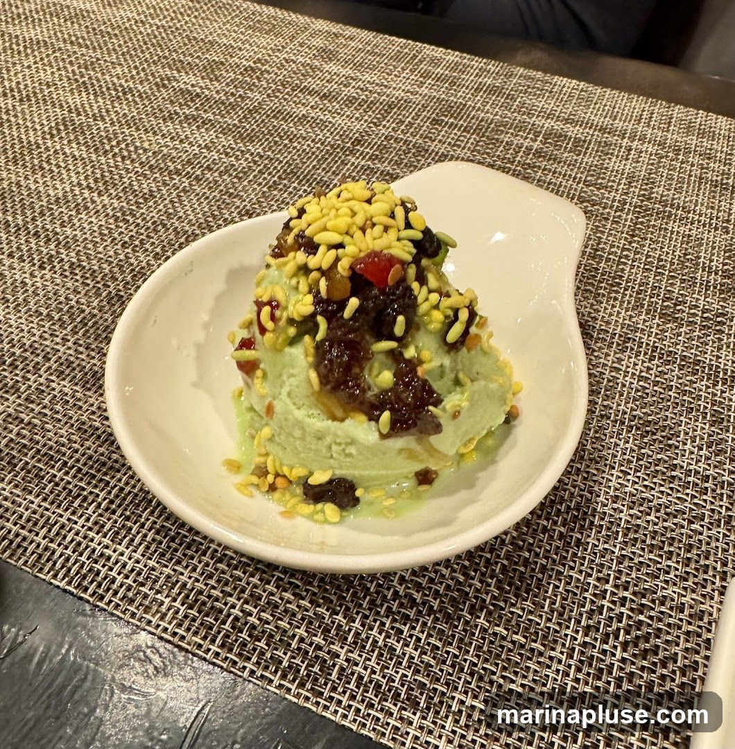 A scoop of light green pistachio ice cream is served in a white, curved dish. The ice cream is topped with a dark, jam-like substance, dried fruits such as raisins, and small, yellow puffed grains. The dish sits on a textured, brown placemat.