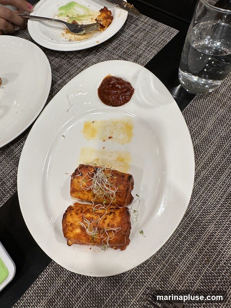 Two pieces of paneer tikka, a popular Indian appetizer, are served on a white plate with a dollop of chutney and garnished with sprouts. The paneer appears to be marinated in a reddish spice blend and grilled or pan-fried. The plate is partially eaten, with some residual oil and sauce visible. A glass of water and another plate with remnants of food are also on the table.