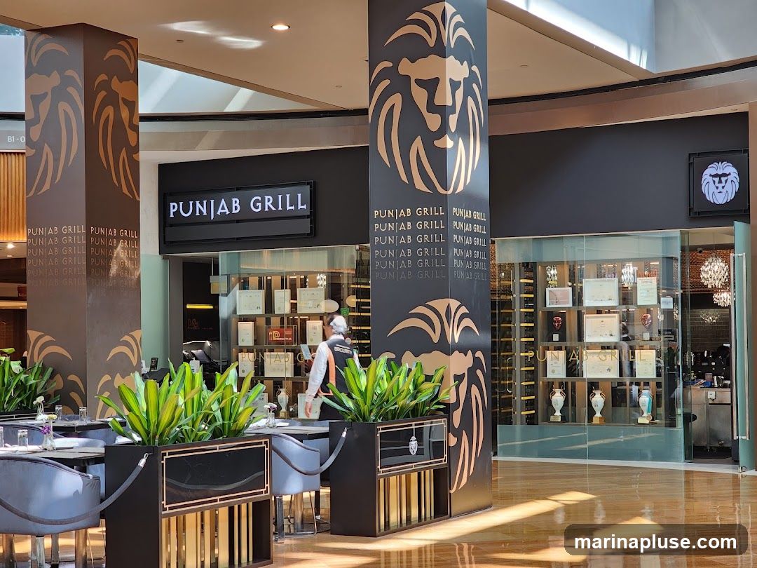 The image shows the interior of a restaurant called Punjab Grill. Large pillars with a lion logo and the restaurant name are visible. The dining area has tables and chairs, and a display case with awards and bottles is seen behind glass. A staff member in uniform is also visible.