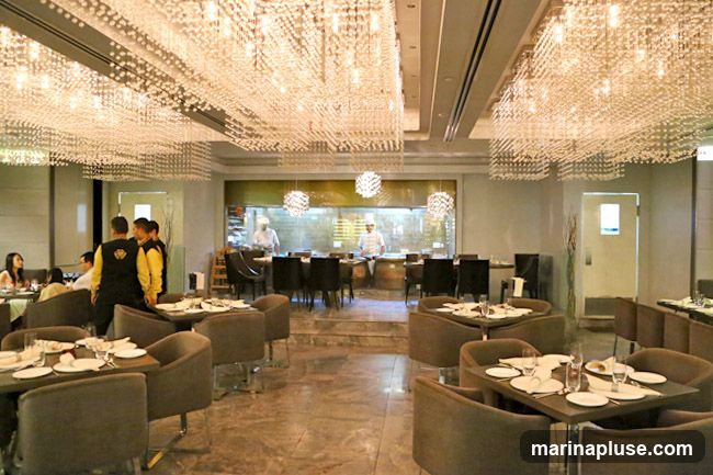The image shows the interior of a restaurant with a view into an open kitchen. The dining area features tables and chairs, with several patrons seated. Waitstaff in uniform are visible. The ceiling is adorned with elaborate crystal chandeliers. In the kitchen, chefs in white uniforms are working.