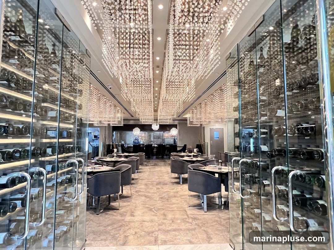 The image shows the interior of a sophisticated restaurant. Floor-to-ceiling glass walls on the left and right display numerous bottles of wine on shelves. The ceiling is adorned with elaborate crystal chandeliers that cast a warm glow. The dining area features tables set with chairs, and in the background, a bar area is visible with staff members. The overall ambiance is luxurious and inviting.