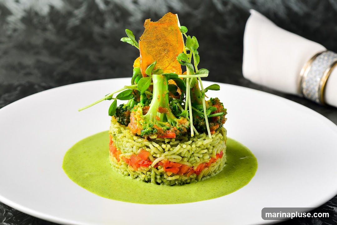 A meticulously plated dish featuring a cylindrical tower of green-tinted rice interspersed with diced red vegetables, topped with broccoli florets, microgreens, and a crispy orange garnish. The dish is served on a white plate with a vibrant green sauce pooled around its base. A white napkin with a decorative ring is visible in the background.