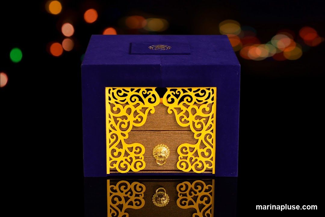 A dark blue velvet box with intricate gold filigree on the front. A golden lion head ornament is centered on the front panel. The box is set against a dark background with blurred colorful lights.