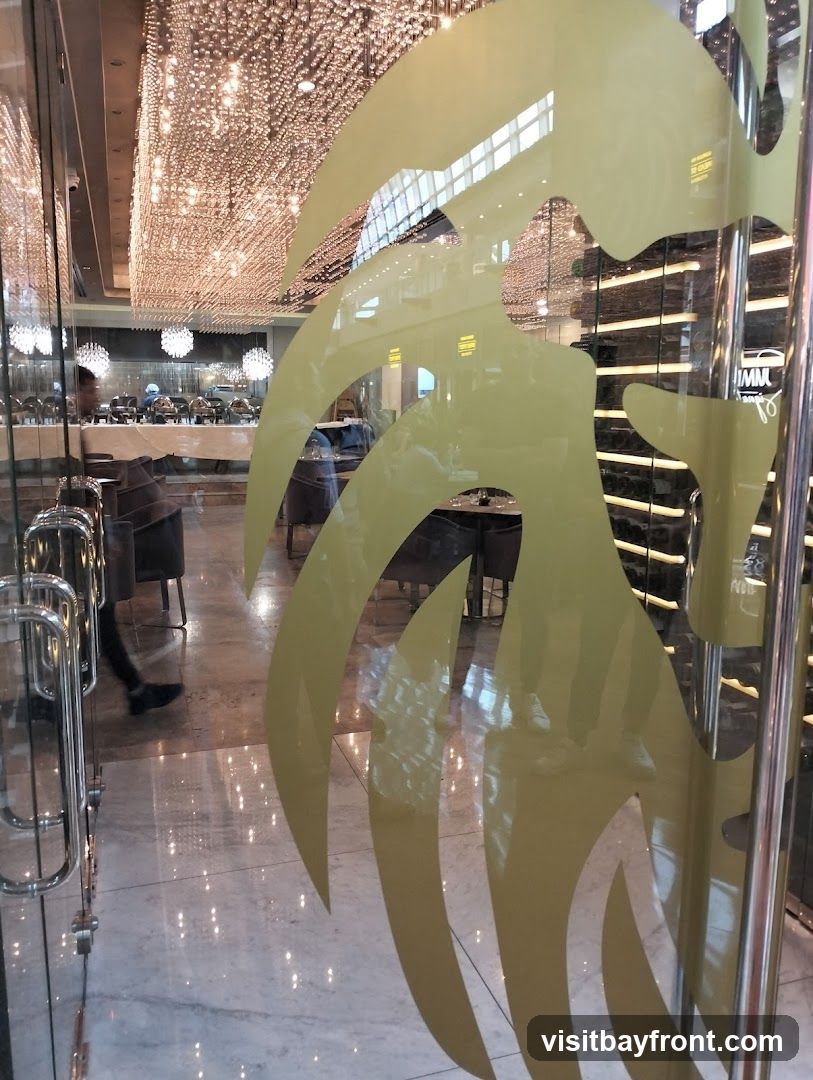 View through a glass door into a restaurant. The interior features elegant decor with numerous hanging lights, a buffet setup with chafing dishes, and seating areas with upholstered chairs and tables. A person is walking into the restaurant. The glass door has a decorative yellow pattern.