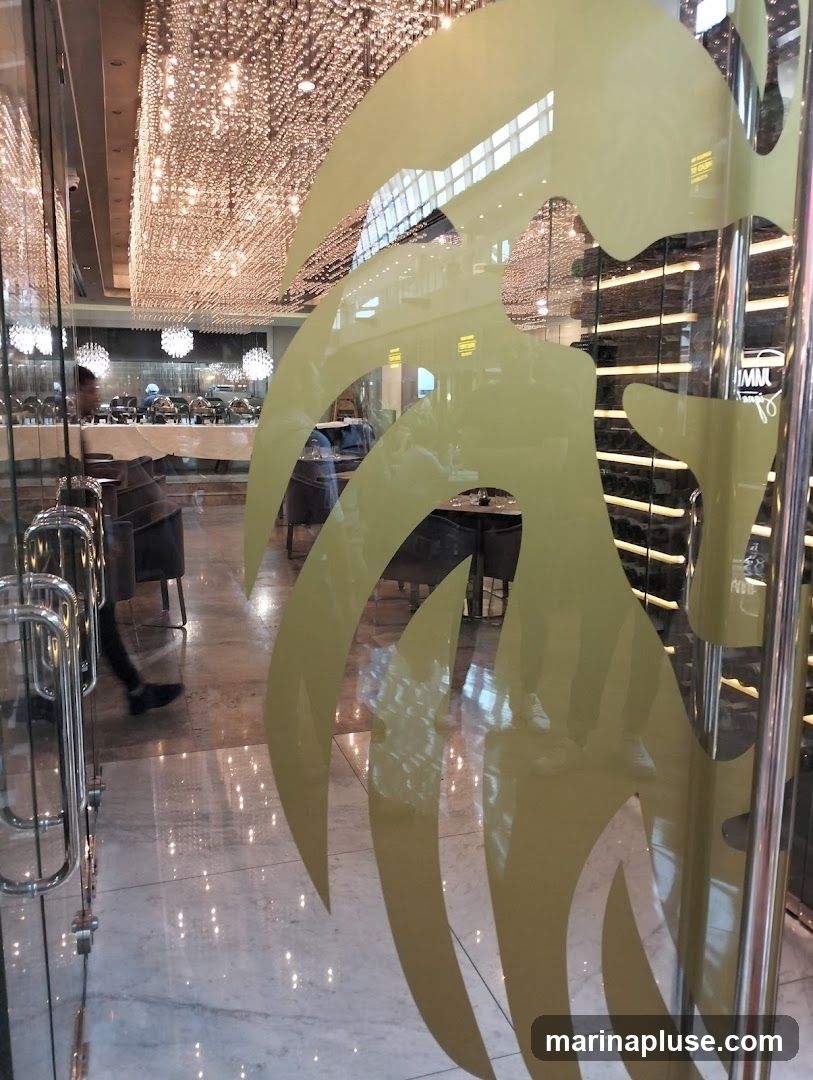 View through a glass door into a restaurant. The interior features elegant decor with numerous hanging lights, a buffet setup with chafing dishes, and seating areas with upholstered chairs and tables. A person is walking into the restaurant. The glass door has a decorative yellow pattern.