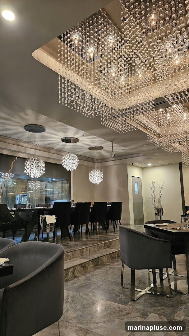 The image shows the interior of a luxurious restaurant. Several large, ornate chandeliers made of crystal beads hang from the ceiling, casting a warm glow. The dining area features dark, plush chairs and tables set with white linens. A raised section of the floor is visible, leading to a long dining table. In the background, a glimpse of the kitchen area can be seen through a glass partition. The overall ambiance is sophisticated and upscale.